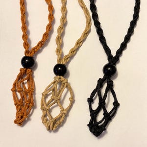 May include: Three macrame necklaces in shades of orange, tan, and black. Each necklace features a woven pouch with a black bead at the top. The cords are braided, and the pouches are designed to hold small items.