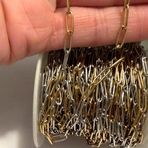 Può includere: Una bobina di catena in metallo misto, con maglie alternate oro e argento. La catena è tenuta in alto, mostrando il contrasto tra i due colori. Le maglie sono di forma ovale e la catena viene probabilmente utilizzata per la creazione di gioielli o lavori artigianali.