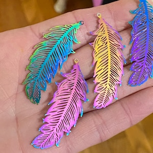 May include: Four iridescent metal feather charms featuring intricate cut-out details. Each charm displays a vibrant gradient of colors, including blue, green, yellow, orange, and purple, and includes a small circular loop at the top for attachment.
