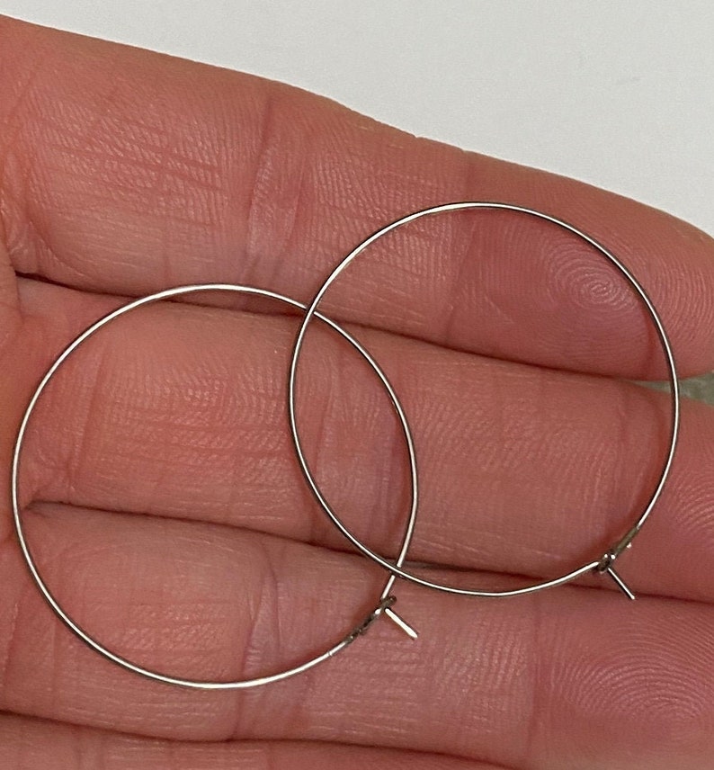 Op de afbeelding: Een paar dunne, cirkelvormige zilverkleurige oorringen. Elke oorbel heeft een eenvoudig, doorlopend draadontwerp met een klein, ge&iuml;ntegreerd sluitingsmechanisme waar de twee uiteinden samenkomen, wat zorgt voor een strak en eenvoudig uiterlijk.