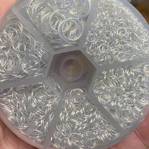 May include: A clear, segmented container filled with various sizes of silver-colored jump rings. The jump rings are neatly organized, with different compartments holding varying quantities and sizes. Ideal for jewelry making and crafting.
