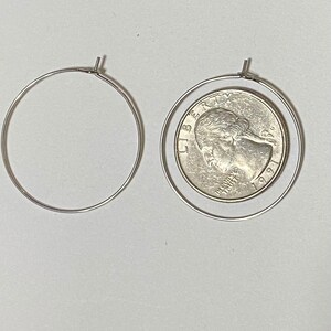 Op de afbeelding: Twee dunne zilverkleurige draad-oorringen. De ene ring is leeg, terwijl de andere ring een Amerikaanse kwartdollar uit 1991 omsluit, met het profiel van George Washington en de tekst LIBERTY en IN GOD WE TRUST.