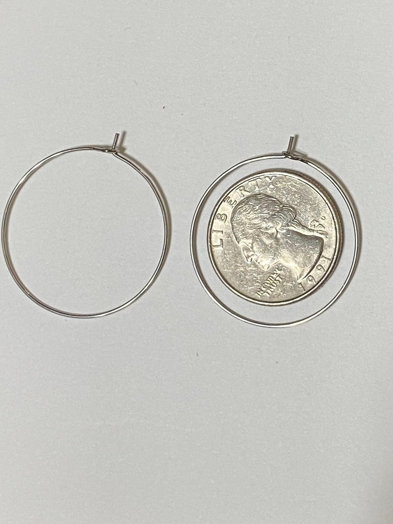 Op de afbeelding: Twee dunne zilverkleurige draad-oorringen. De ene ring is leeg, terwijl de andere ring een Amerikaanse kwartdollar uit 1991 omsluit, met het profiel van George Washington en de tekst LIBERTY en IN GOD WE TRUST.