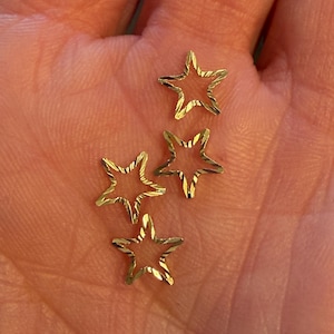 May include: Four small, gold-colored star-shaped charms with an open center. Each star features a textured, diamond-cut surface that creates a reflective, shimmering effect. The stars are arranged in a cluster, showcasing their metallic finish.