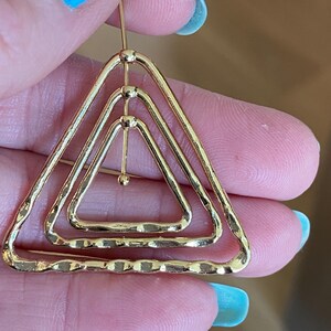 May include: A gold-toned earring featuring three nested triangular frames with a hammered, textured surface. The triangles are connected by a vertical central bar with small spherical accents at each junction, creating a layered geometric design.