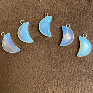 May include: Five iridescent opal crescent moon charms with silver jump rings. The charms are a light blue color with a pearly sheen.