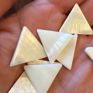 May include: A collection of small, white, triangular decorative pieces with a pearlescent sheen. The pieces are smooth and have rounded edges. They are held in a hand, suggesting their small size and delicate nature.