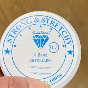 May include: A white round container with blue text that reads "Strong & Stretchy" with a diamond logo and the text "0.7 水晶の線 CRYSTALLINE Size: Color: 100%"