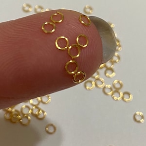 May include: A close-up of a fingertip holding a cluster of small, gold jump rings. The rings are scattered on a white surface.