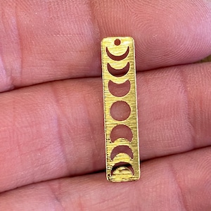 May include: Gold-colored metal charm with a cutout design of the phases of the moon. The charm is rectangular and has a textured surface.