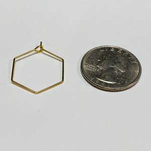 Puede incluir: Un pendiente hexagonal dorado junto a una moneda de veinticinco centavos. El pendiente tiene una forma geom&eacute;trica simple, y la moneda sirve como referencia de tama&ntilde;o. El pendiente est&aacute; hecho de alambre de metal fino.