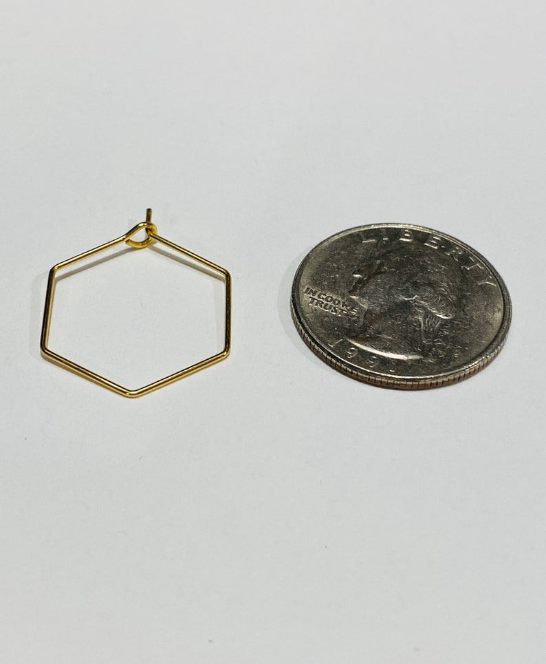 Puede incluir: Un pendiente hexagonal dorado junto a una moneda de veinticinco centavos. El pendiente tiene una forma geom&eacute;trica simple, y la moneda sirve como referencia de tama&ntilde;o. El pendiente est&aacute; hecho de alambre de metal fino.