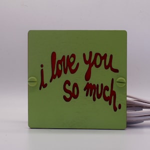 May include: A green wooden sign with the words "i love you so much." written in red.