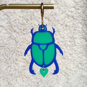 Peut inclure: Boucle d'oreille en forme de scarabée bleu et vert. La boucle d'oreille présente un contour bleu avec un centre vert et un petit pendentif en forme de cœur vert. La boucle d'oreille est suspendue à un anneau doré.