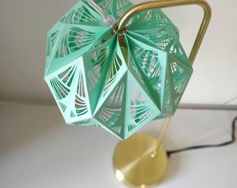 Könnte beinhalten: Eine goldene Tischlampe mit einem grünen Papierschirm. Der Schirm hat ein geometrisches Design mit ausgeschnittenen Details.
