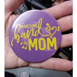 May include: A round, purple button with the words "MARCHING band MOM" in yellow script and block letters. Musical notes and a treble clef are also featured in yellow. The button is held in a hand.