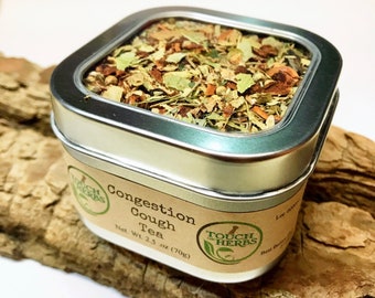May include: A square, silver tin of "Congestion Cough Tea" by Touch of Herbs, filled with dried herbs and leaves. The label indicates a net weight of 2.5 oz (70g). The tin sits on a piece of wood against a white background.