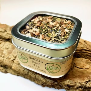 Peut inclure: Une bo&icirc;te carr&eacute;e &agrave; bords argent&eacute;s remplie de th&eacute; en vrac, &eacute;tiquet&eacute;e "Healthy Head Tea". L'&eacute;tiquette indique "Touch of Herbs" et le poids net est de 50g. La bo&icirc;te est pos&eacute;e sur un morceau de bois flott&eacute;.