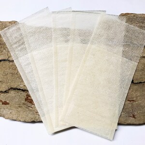 Peut inclure: Cinq sachets de th&eacute; rectangulaires, de couleur blanc cass&eacute;, fabriqu&eacute;s &agrave; partir d'un mat&eacute;riau fin et textur&eacute;. Ils sont dispos&eacute;s en &eacute;ventail. L'arri&egrave;re-plan est un morceau de bois brun clair vieilli avec des fissures sombres.
