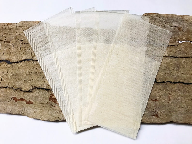 Peut inclure: Cinq sachets de th&eacute; rectangulaires, de couleur blanc cass&eacute;, fabriqu&eacute;s &agrave; partir d'un mat&eacute;riau fin et textur&eacute;. Ils sont dispos&eacute;s en &eacute;ventail. L'arri&egrave;re-plan est un morceau de bois brun clair vieilli avec des fissures sombres.