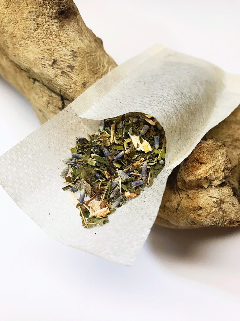 Peut inclure: Un sachet de th&eacute; blanc rempli d'un m&eacute;lange d'herbes et de fleurs s&eacute;ch&eacute;es, dont de la lavande et de la menthe. Le sachet de th&eacute; est ouvert, r&eacute;v&eacute;lant le contenu color&eacute;. Le sachet de th&eacute; repose sur un morceau de bois flott&eacute; sur fond blanc.