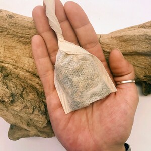 Peut inclure: Un sachet de th&eacute; carr&eacute; rempli de th&eacute; en vrac, tenu dans une main ouverte. Le sachet de th&eacute; est fait d'un papier clair et textur&eacute; et a un dessus torsad&eacute;. La main est orn&eacute;e d'une bague en argent et d'un bracelet en argent &agrave; motifs. L'arri&egrave;re-plan comprend un morceau de bois flott&eacute;.