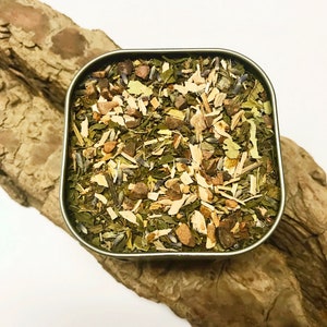 Peut inclure: Une bo&icirc;te carr&eacute;e en argent remplie d'un m&eacute;lange d'herbes et de plantes s&eacute;ch&eacute;es, comprenant des feuilles vertes, de la lavande et des copeaux de bois. La bo&icirc;te est pos&eacute;e sur un morceau de bois brun clair textur&eacute;.