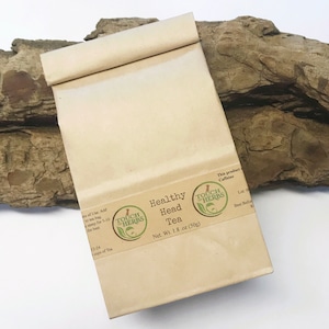 Peut inclure: Un sachet en papier beige de "Healthy Head Tea" avec une &eacute;tiquette marron et un logo vert. L'&eacute;tiquette indique "Net. Wt. 50g". Le sachet est pos&eacute; sur un morceau de bois flott&eacute;.