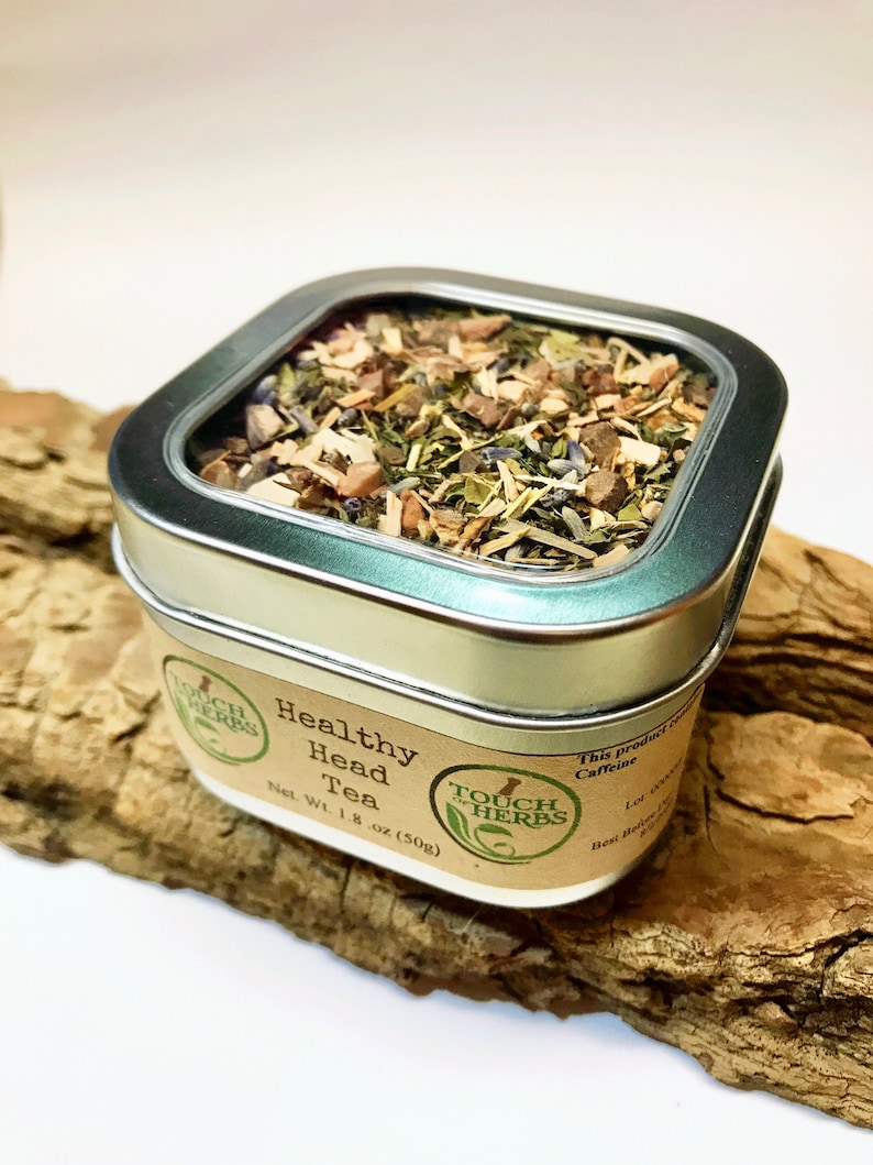 Peut inclure: Une bo&icirc;te carr&eacute;e &agrave; bords argent&eacute;s remplie de th&eacute; en vrac, &eacute;tiquet&eacute;e "Healthy Head Tea". L'&eacute;tiquette indique "Touch of Herbs" et le poids net est de 50g. La bo&icirc;te est pos&eacute;e sur un morceau de bois flott&eacute;.
