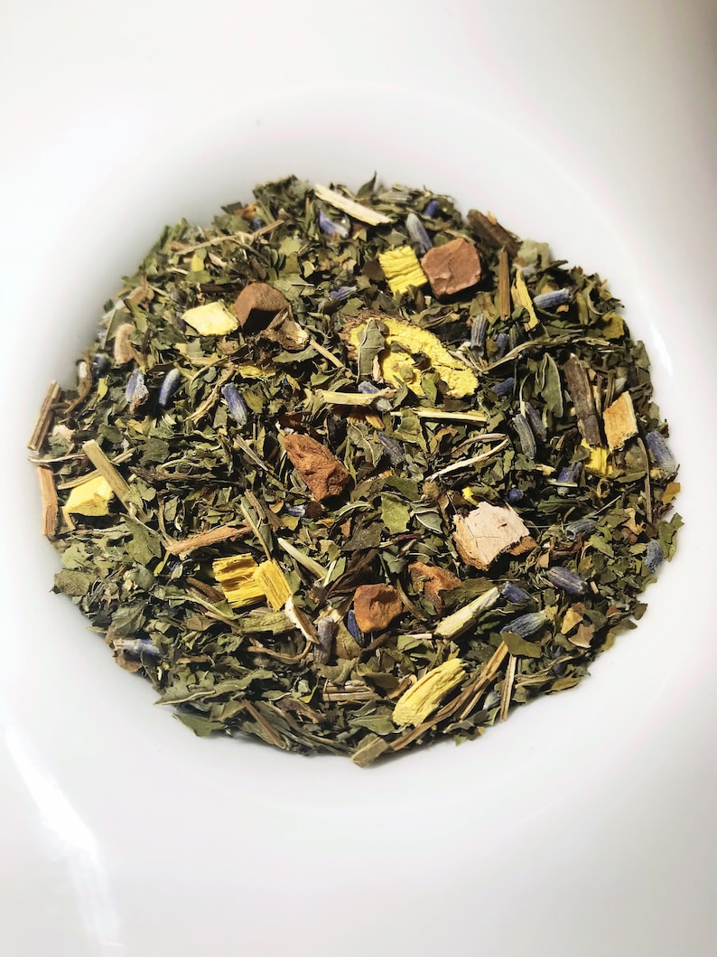Peut inclure: Gros plan d'un m&eacute;lange de th&eacute; de plantes en vrac dans un bol blanc. Le th&eacute; contient des feuilles vertes, des boutons de lavande violets et des morceaux d'herbes jaunes et brunes. Le m&eacute;lange de th&eacute; pr&eacute;sente une vari&eacute;t&eacute; de textures et de couleurs.