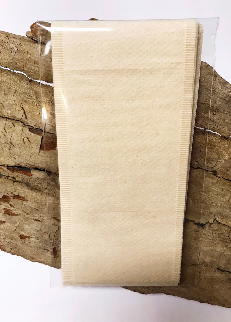 Peut inclure: Un paquet de filtres &agrave; th&eacute; beiges dans un emballage plastique transparent. Les filtres sont rectangulaires et ont une surface textur&eacute;e. L'emballage repose sur un morceau de bois vieilli.