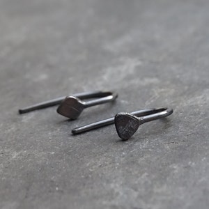 May include: A pair of dark grey metal earrings. Each earring has a small, textured, geometric shape connected to a curved bar and a straight post. The earrings are set against a grey background.