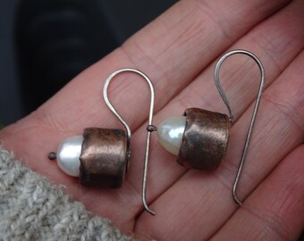 May include: A pair of handcrafted earrings featuring white pearl beads encased in copper cylinders. The copper has a textured, aged appearance. The earrings have silver-colored, curved ear wires.