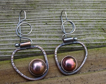 May include: A pair of handcrafted earrings with a unique design. Each earring features a hammered copper frame with a central copper sphere. The ear wires and upper decorative elements are made of silver-colored metal. The earrings are approximately 2 inches long.