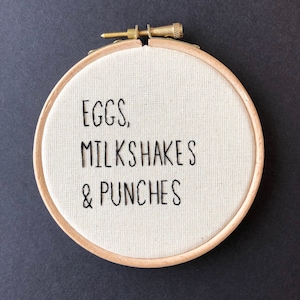 May include: A white embroidery hoop with the text "EGGS, MILKSHAKES & PUNCHES" embroidered in black thread.