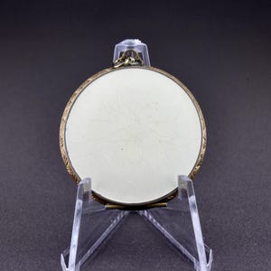 Op de afbeelding: Een rond, wit porseleinen medaillon met een goudkleurig metalen frame. Het medaillon heeft een delicaat bloemmotief dat in het porselein is gegraveerd. Het medaillon staat op een transparante kunststof standaard.
