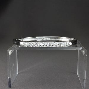 May include: A silver hinged bangle bracelet, set with small, clear crystals. The bracelet is displayed on a clear acrylic stand against a dark grey background. The crystals sparkle, reflecting light.