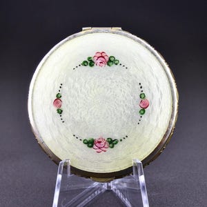 May include: A round, white compact with a silver rim. The top of the compact is decorated with a painted design of pink roses and green leaves.