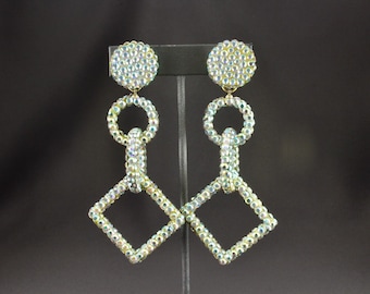 May include: A pair of dazzling, iridescent rhinestone earrings. Each earring features a round stud, a circular link, and a diamond-shaped pendant. The earrings are designed to dangle and catch the light, creating a sparkling effect. The earrings are displayed against a dark background.