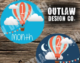 May include: Three round blue and white stickers with hot air balloons and numbers 1, 2, and 3. The balloons are red and white striped with blue accents. The stickers say "month" or "months" and are for baby milestones.