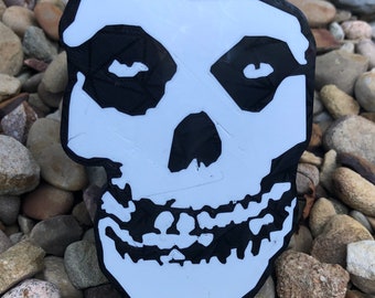 May include: A white skull with black eye sockets and a black smile. The skull is made of plastic and has a 3D effect.