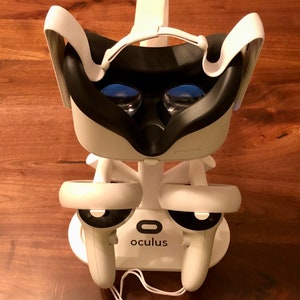 Puede incluir: Auriculares VR Oculus blancos con dos mandos blancos en un soporte blanco. El soporte tiene la palabra "oculus" impresa.