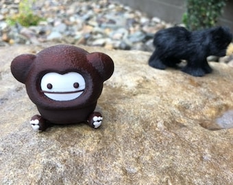 May include: A brown monkey figurine with a white face and small white feet. A black bear figurine is in the background. Both figurines are on a large, textured rock.