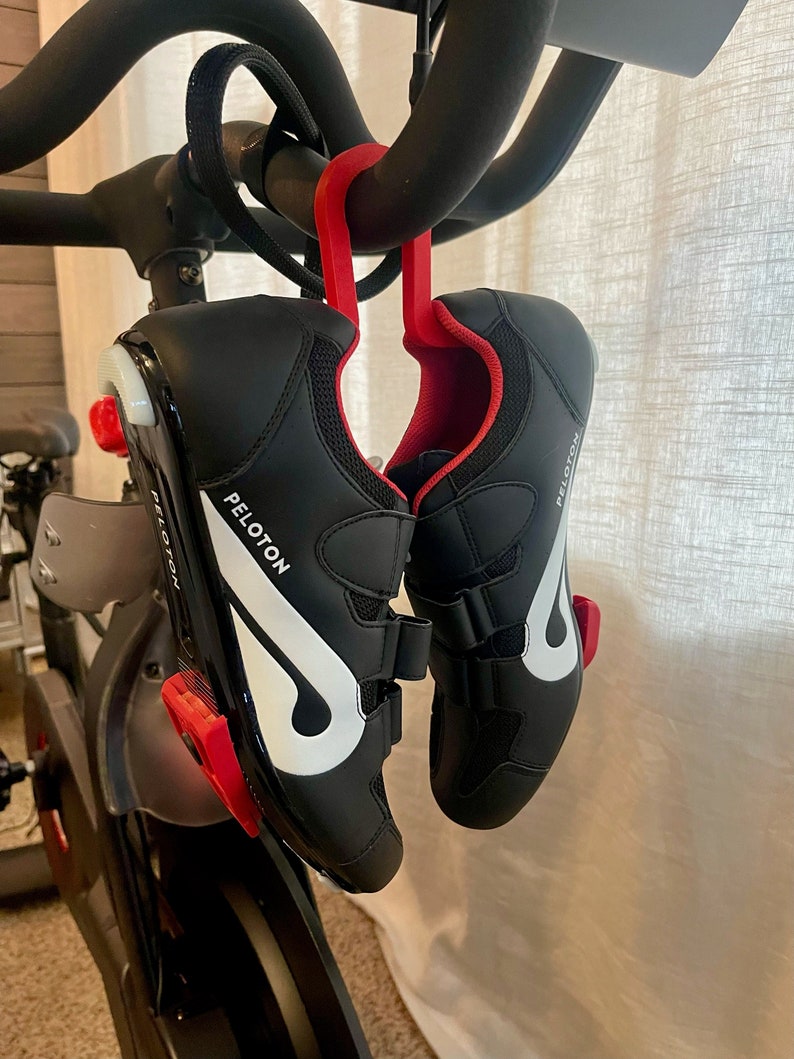 Puede incluir: Un par de zapatillas de ciclismo negras con logotipos Peloton blancos colgados de un gancho rojo sujeto a una bicicleta de ejercicio negra.