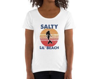 May include: White scoop neck t-shirt with a graphic design. The design features a silhouette of a mermaid inside a sunset circle with the words "SALTY" and "LIL' BEACH" in blue text. The shirt is made of a soft material.