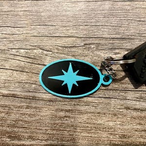 May include: A black oval keychain with a blue starburst design. The keychain is attached to a silver key ring and a black key.