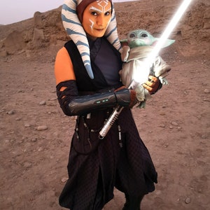 May include: A woman dressed as Ahsoka Tano from Star Wars, wearing a blue and white headpiece, a black and brown outfit, and holding a lightsaber. She is holding a small green alien doll in her other hand.