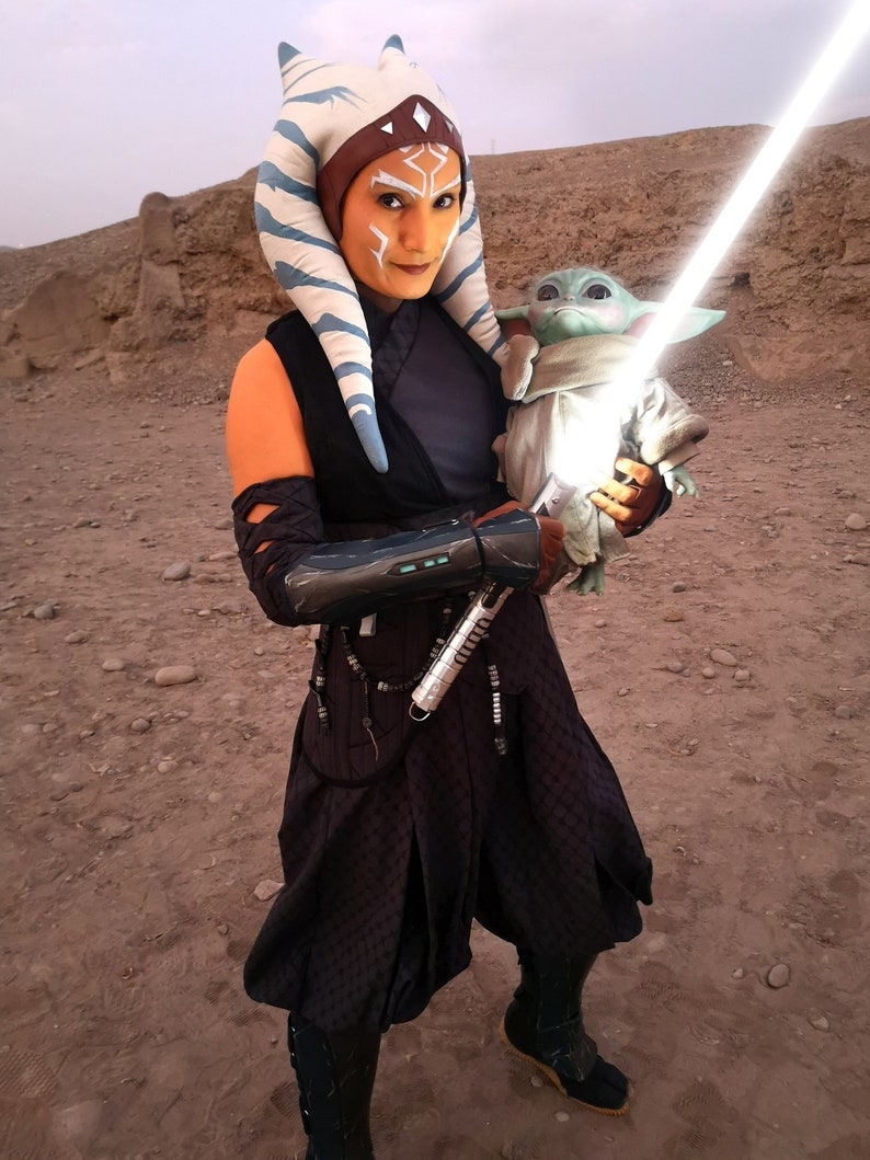 May include: A woman dressed as Ahsoka Tano from Star Wars, wearing a blue and white headpiece, a black and brown outfit, and holding a lightsaber. She is holding a small green alien doll in her other hand.