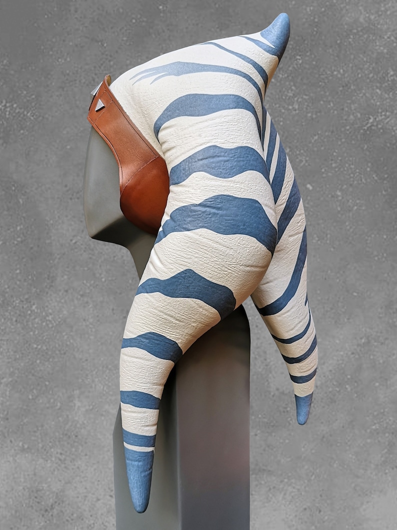 May include: A white and blue striped headpiece with a brown leather strap. The headpiece is shaped like two horns and is designed to resemble a Twi'lek head.