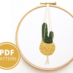 Op de afbeelding: Een groene cactusplant in een gele hangende pot met een witte kwast. De plant bevindt zich in een houten borduurring.